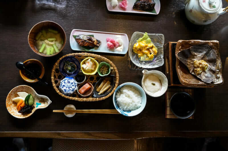 Japanese ryokan breakfast dishes including cooked white rice, grilled fish, fried egg, soup, mentaiko, pickle, seaweed, hot plate, other side dishes and green tea on wooden table, Japan = Sgutterstock